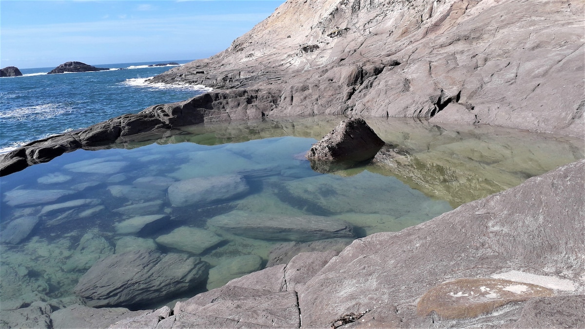 A rocky coastline features a clear tidal pool reflecting the sky. Nearby, the ocean waves gently lap against the rugged rocks, while distant landforms create a scenic backdrop. The varied textures of the rock formations are visible, highlighting the area's natural beauty.