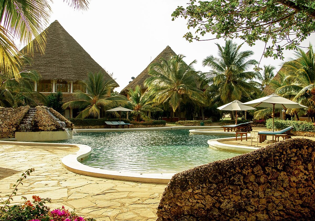A tranquil pool area is surrounded by lush palm trees and thatched-roof structures. The pool features a gentle waterfall, and lounge chairs are positioned under large umbrellas, providing shaded spaces for relaxation. The stone walkway leads toward the water's edge.