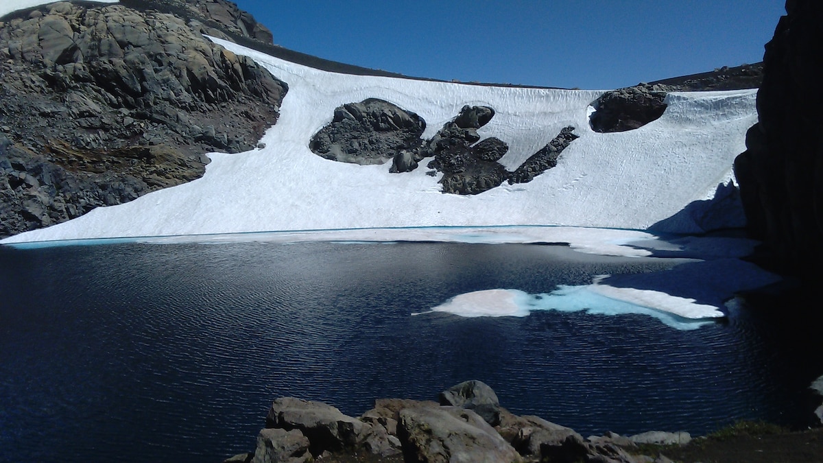 A serene glacial lake is framed by rocky terrain, with snow-covered mountains in the background. The clear blue water reflects the surrounding landscape. Ice patches float gently on the surface, providing a contrast to the deep tones of the lake, under a clear blue sky.
