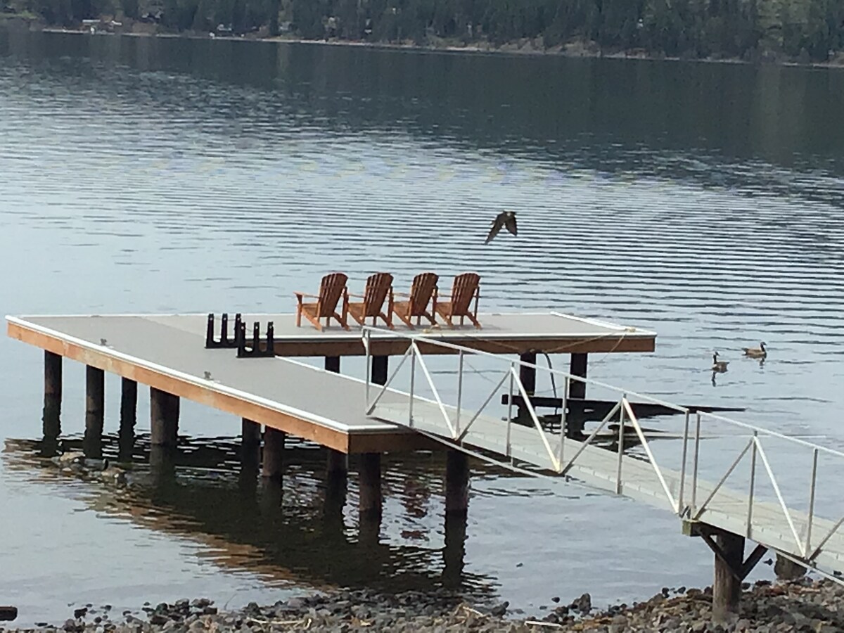A wooden dock extends into the calm lake water, featuring three wooden lounge chairs facing the serene landscape. The surface reflects the surrounding trees and gentle ripples, while a few ducks can be spotted nearby, contributing to the tranquil setting.