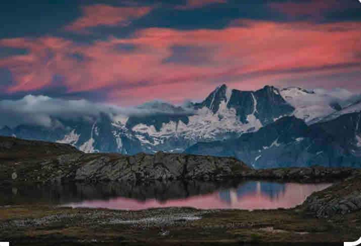 A serene mountain landscape features jagged peaks partially covered in snow, reflecting muted pink and purple hues from the sunset in a tranquil lake. The foreground consists of rugged rock formations, enhancing the natural beauty of the scene.
