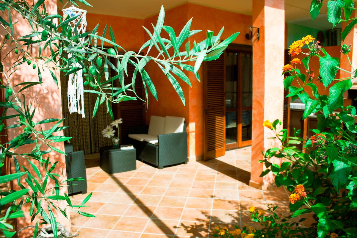 The spacious outdoor area features a comfortable seating arrangement under a covered patio, surrounded by lush greenery and colorful flowers. The tiled floor adds a natural touch, while the warm orange walls enhance the inviting atmosphere.