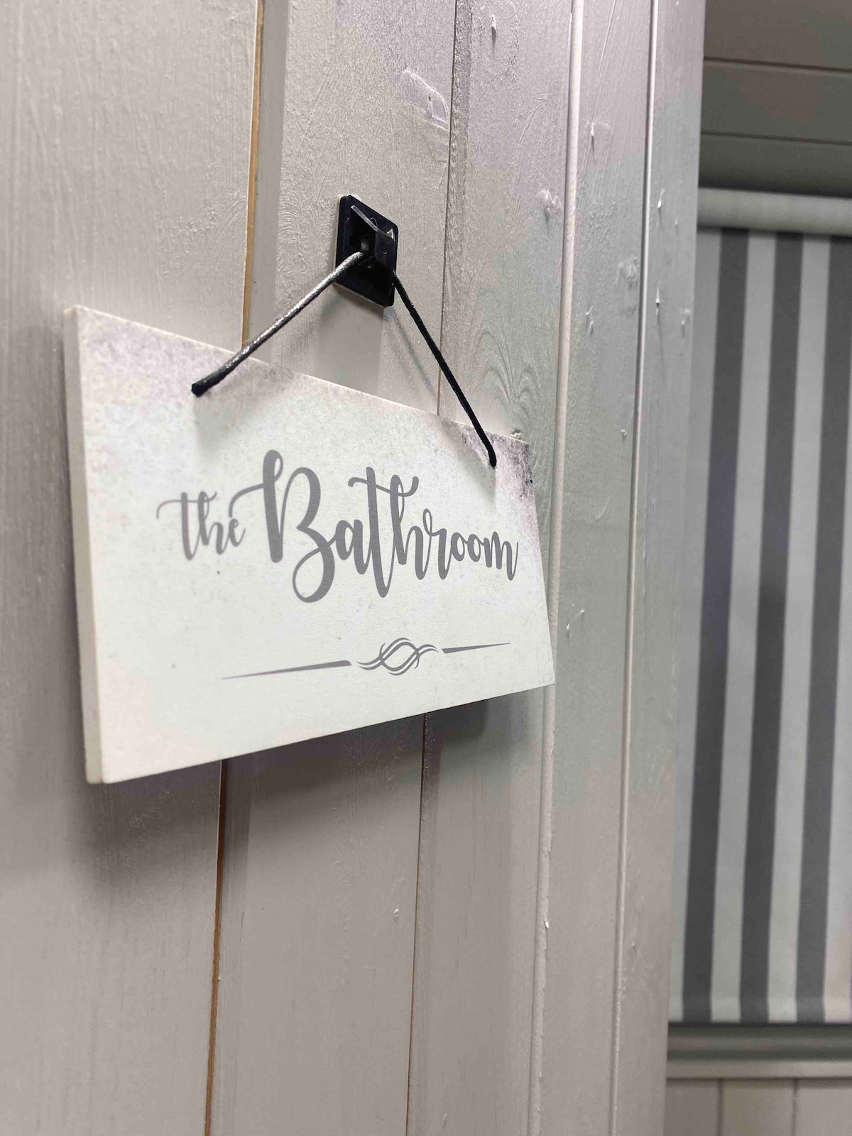 A sign labeled 'the Bathroom' is hanging on a wooden wall, adding a touch of charm. The background features striped window coverings, subtly enhancing the space's character. The light grey tones of the wall create a calm and neutral environment.