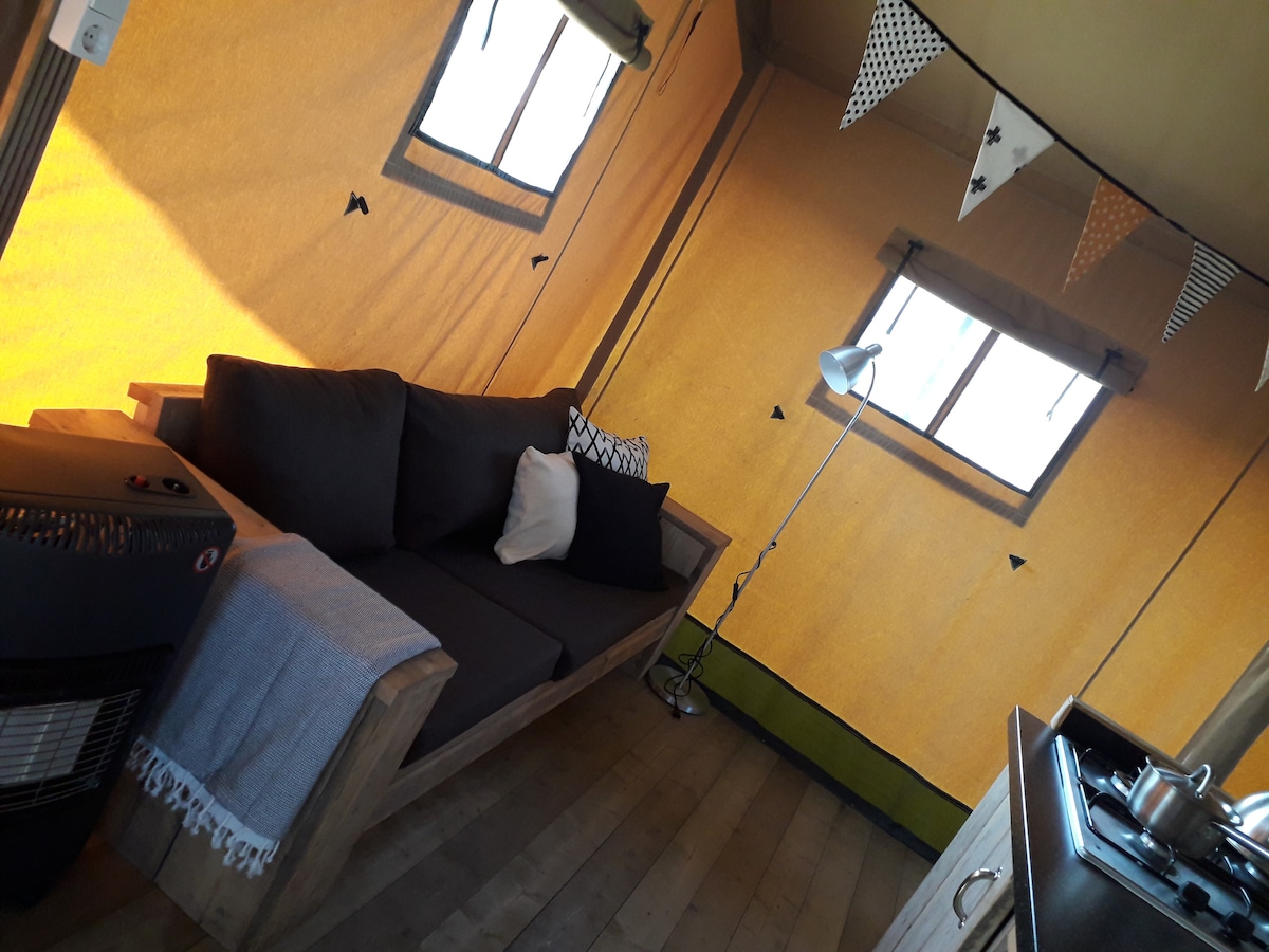 An inviting interior of a cozy safari tent is visible, featuring a comfortable seating area with a dark sofa adorned with decorative cushions. Natural light enters through two small windows, and a stylish floor lamp stands nearby. A glimpse of a compact kitchen can be seen on the right.