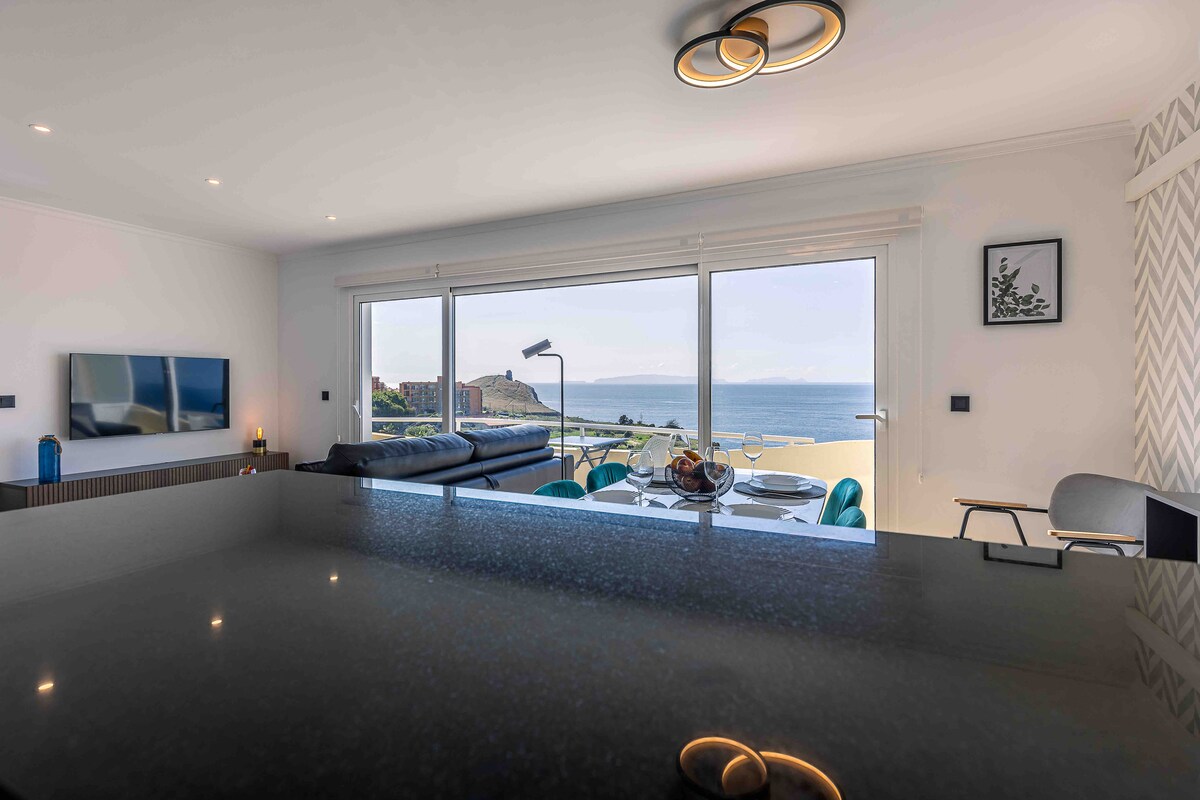 A bright living area is highlighted by large glass doors that open to a stunning ocean view. A modern kitchen counter with sleek surfaces separates the dining space from the lounge area, where a comfortable sofa and stylish table settings are arranged. Natural light streams through, creating an inviting atmosphere.
