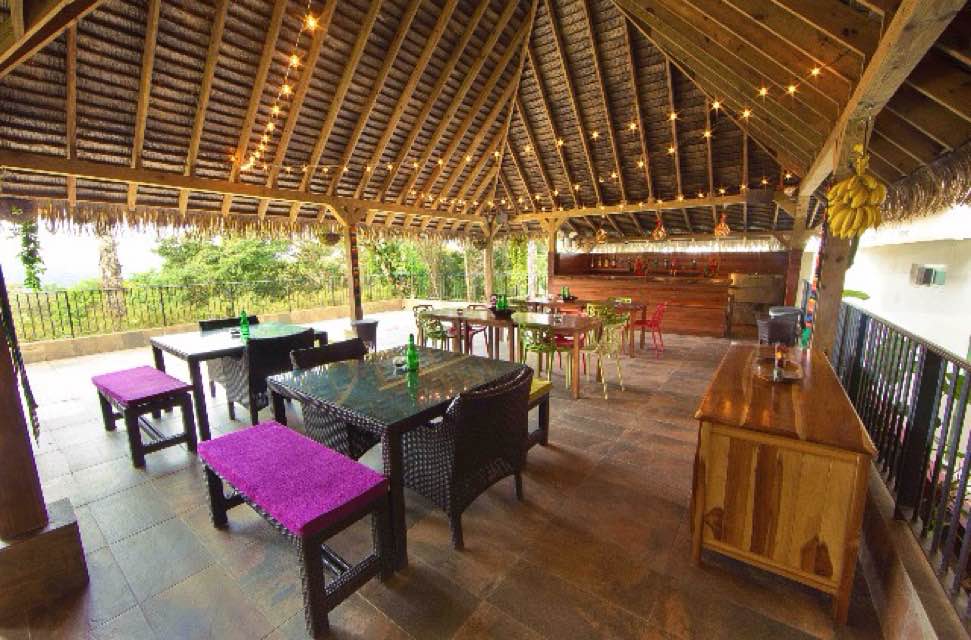 An open-air dining area features wooden beams and a thatched roof. Various tables and chairs are arranged, with two purple benches and a wooden side table visible. String lights illuminate the space, and a lush view of greenery is framed by the railing.
