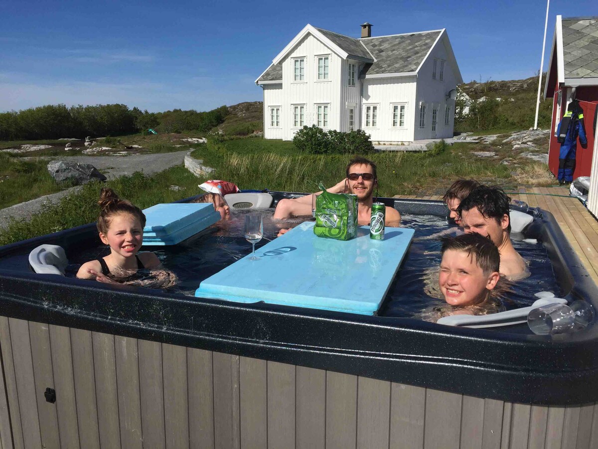 A hot tub is situated on a wooden deck, surrounded by lush greenery. In the background, a traditional coastal house with white siding and a slate roof is visible. Clear skies and natural light create a serene environment.