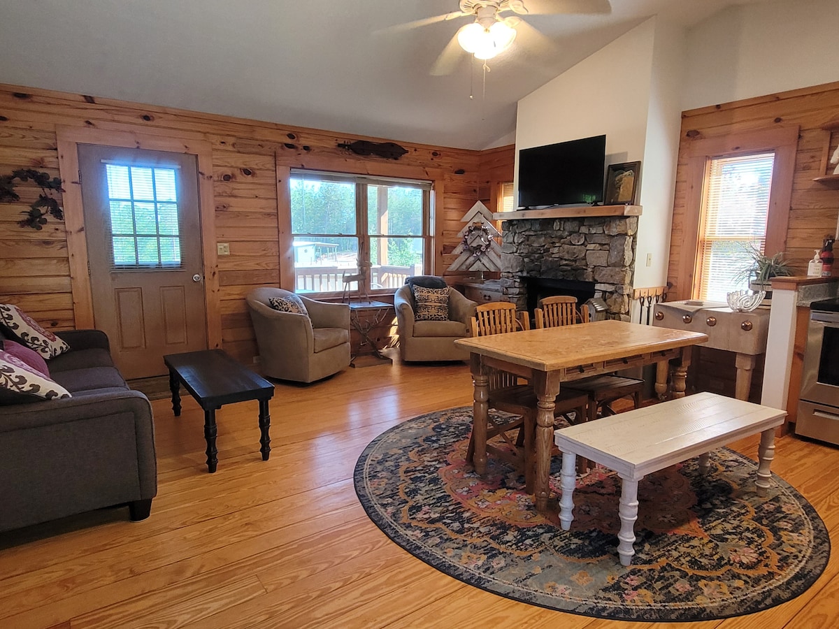 A living area features wooden walls and natural light from large windows. Comfortable seating includes two armchairs and a sofa, arranged around a central wooden table. A rug enhances the space underneath, and a stone fireplace is positioned against one wall.