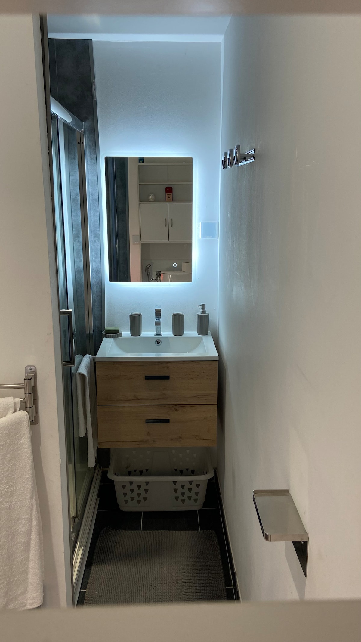 A modern bathroom features a sleek vanity with two basins and a well-lit mirror above. A spacious walk-in shower is visible in the background. White towels hang neatly on a rack, and a laundry basket is placed under the vanity, contributing to the organized and clean appearance.