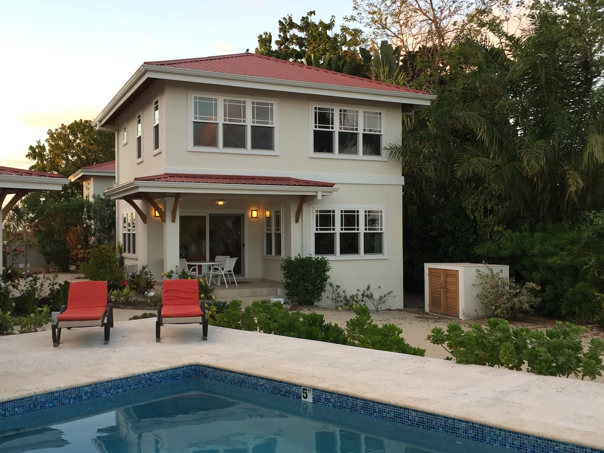 A two-story contemporary house is situated beside a swimming pool. Bright red lounge chairs are positioned near the water, while lush greenery surrounds the property, providing an inviting environment. The house features large windows and a covered porch, emphasizing a seamless connection with the outdoors.