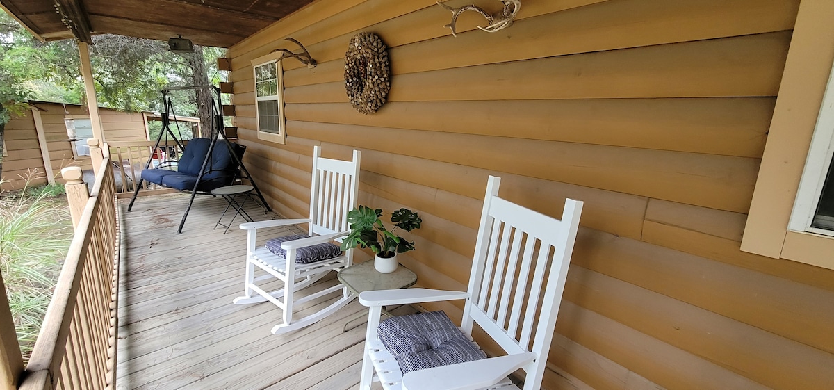 A rustic front porch features two white rocking chairs positioned on a wooden deck. A small potted plant rests on a table between the chairs. The warm wooden siding of the cabin adds to the inviting nature of the outdoor space.