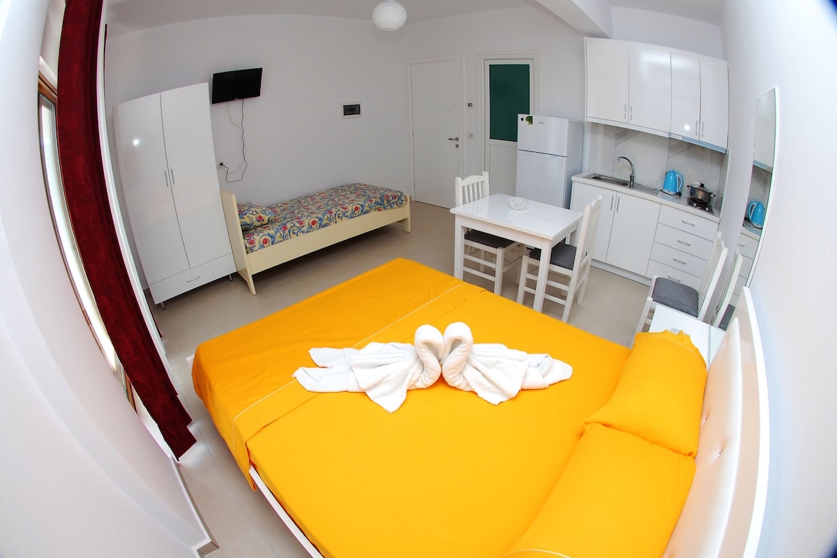 A well-arranged studio features a spacious bed draped in vibrant orange linens, complemented by a colorful patterned single bed. A compact dining table with chairs is positioned nearby, alongside a kitchenette equipped with essential appliances. White cabinetry and neutral walls enhance the overall brightness of the space.