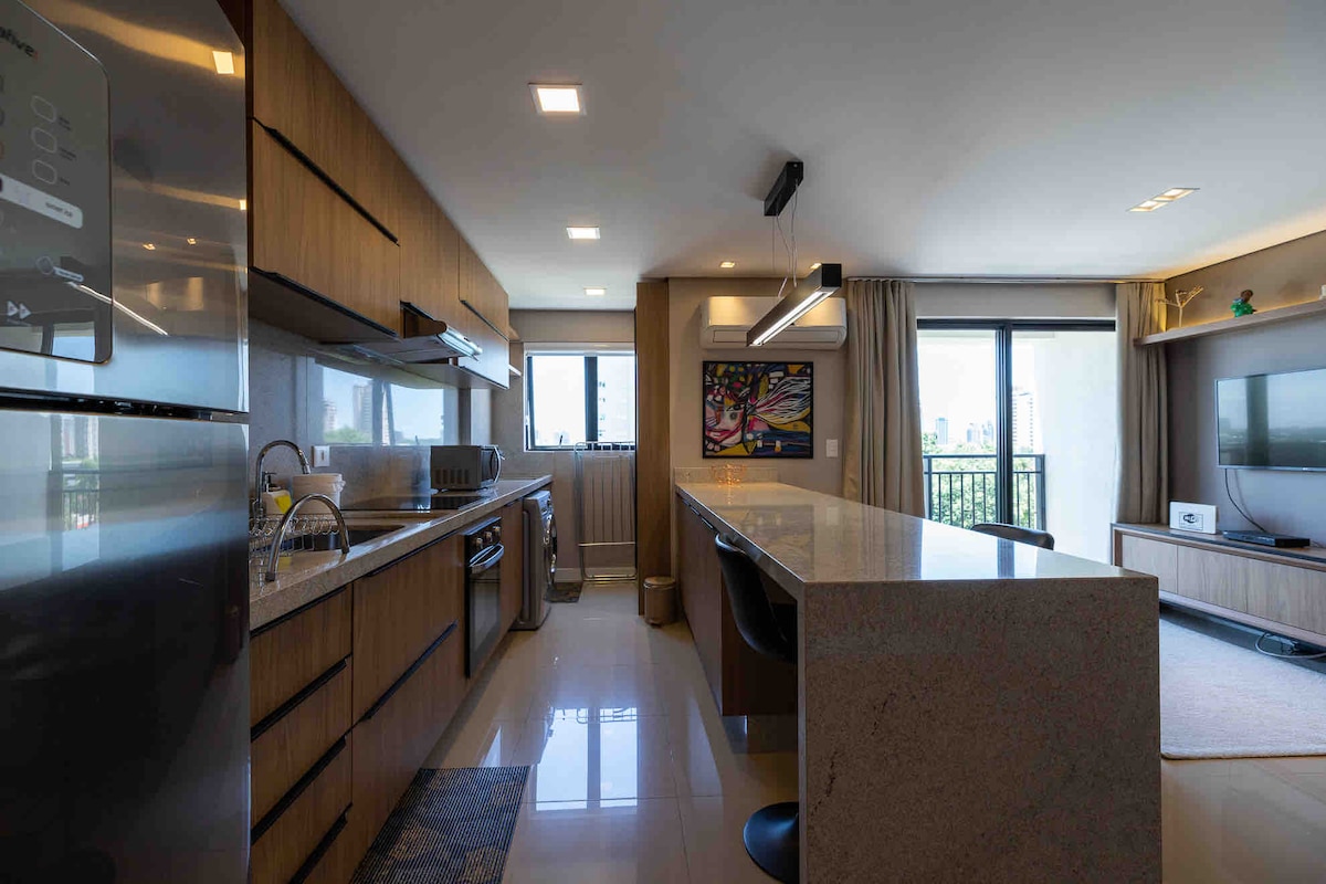 A modern kitchen area features wooden cabinetry and sleek stainless steel appliances. A spacious countertop extends into a dining space with bar stools. A large window allows natural light to fill the room, while a colorful piece of artwork adds a touch of character.