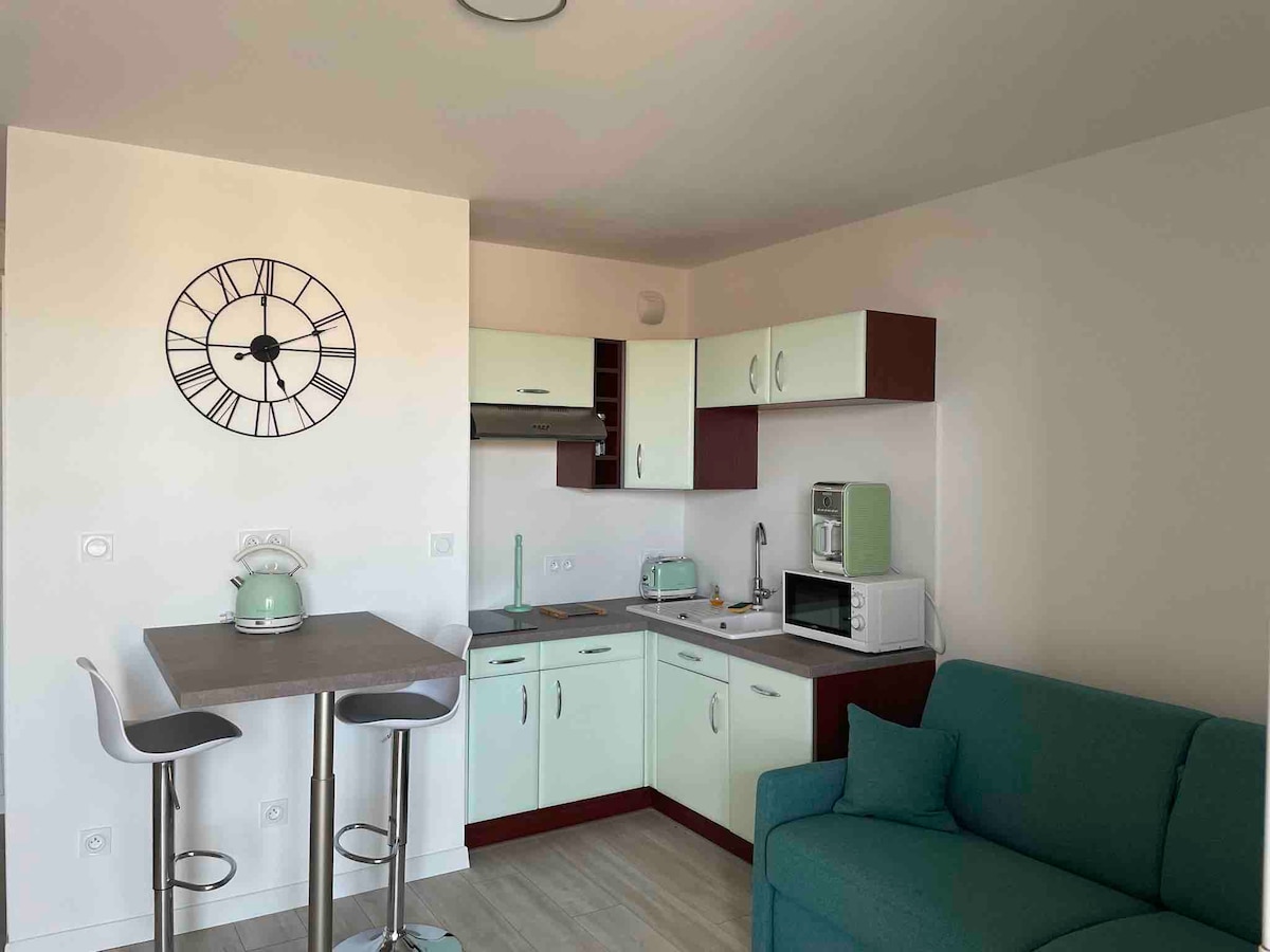 A compact kitchen area is showcased with light-colored cabinetry and modern appliances. A round table with two stools is positioned nearby for dining. A comfortable turquoise sofa is included, providing a cozy seating option in the space.
