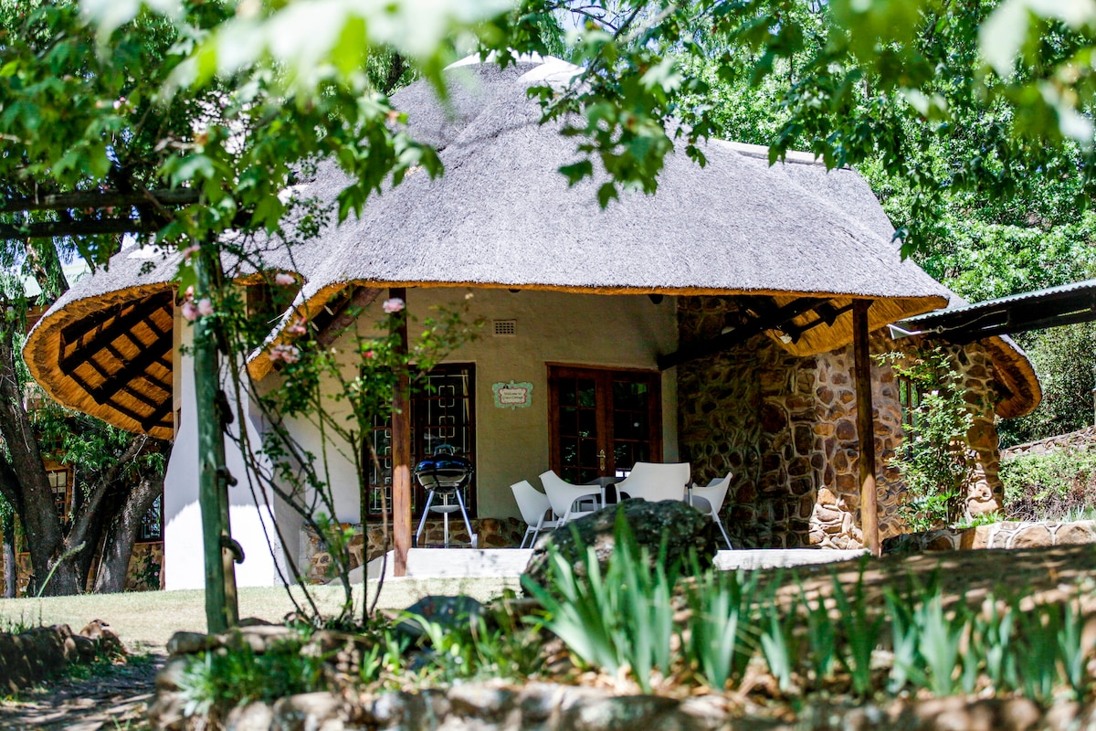 A thatch-roof cottage is nestled among lush greenery, showcasing a stone exterior and wooden accents. The outdoor area features white patio furniture and a barbecue grill, surrounded by vibrant foliage and a peaceful garden setting.