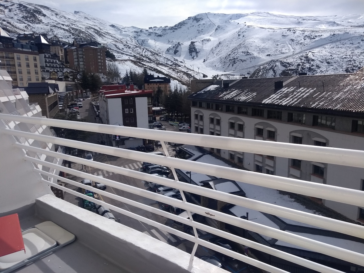 A view from the balcony reveals snow-covered mountains and a bustling street below. Buildings are dotted along the street, with skiing equipment visible. The railing is white, providing a clear frame to the picturesque landscape, capturing the winter atmosphere of Sierra Nevada.