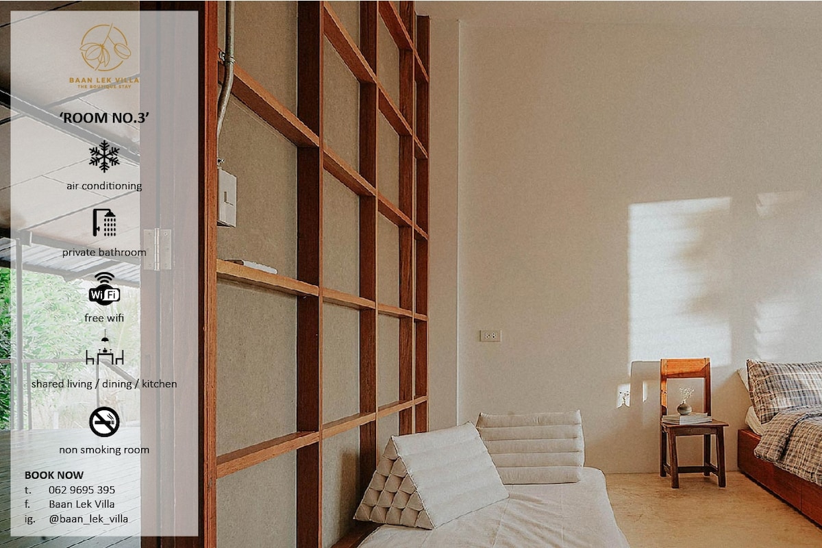 A comfortable guest room features a minimalist design with a high wall displaying wooden shelving. A cozy seating area includes a cushion-covered platform. A wooden chair stands nearby, and a small light illuminates the space, creating a calm environment. Air conditioning and a private bathroom are available.