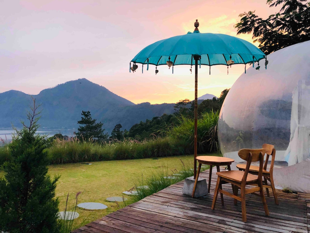 An outdoor seating area features a round teal umbrella providing shade over a small wooden table and two chairs. A lush green lawn extends towards rolling mountains in the background, while a clear, dome-shaped structure is visible nearby, reflecting the soft colors of a sunset sky.