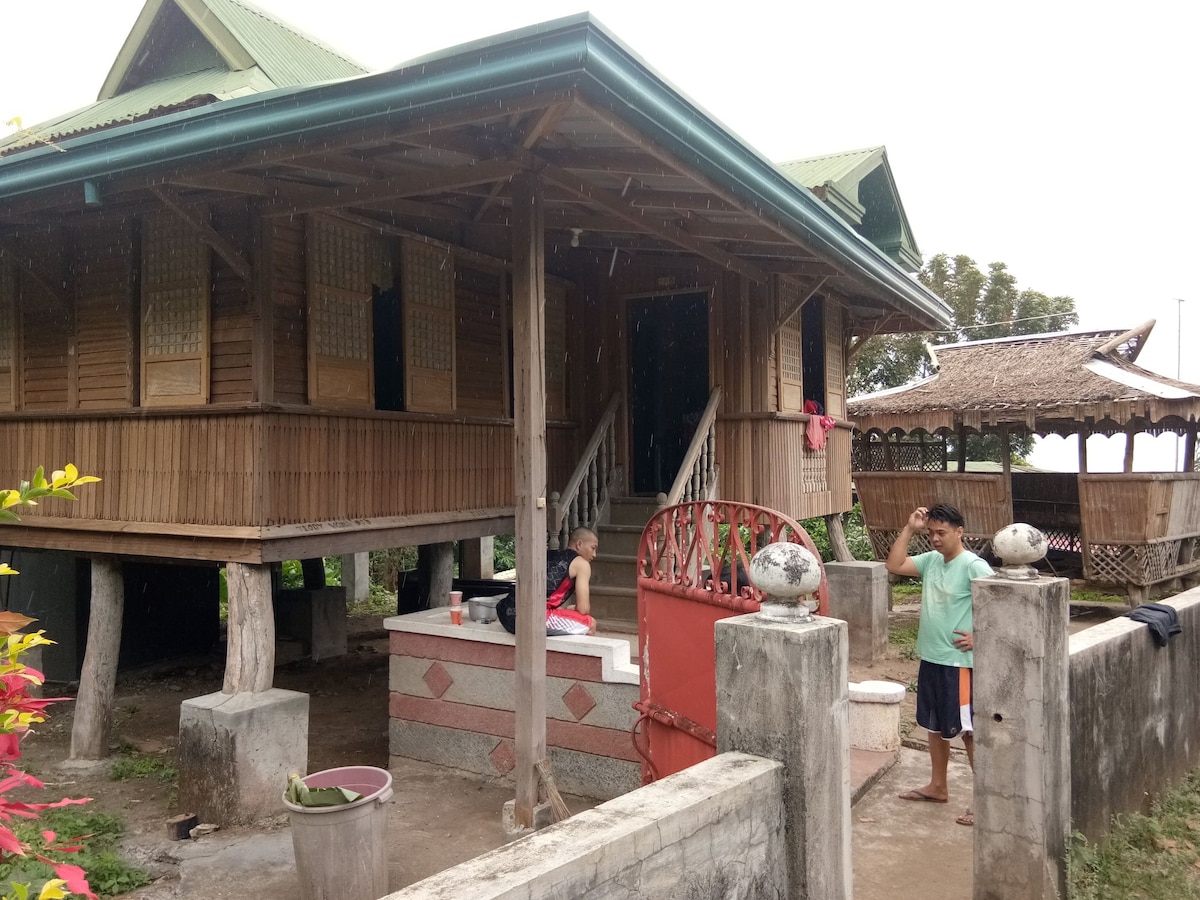 A traditional house is elevated on stilts with a bamboo exterior, surrounded by lush greenery. The structure features multiple wooden windows with woven bamboo screens. A small porch is visible in front, with a couple of people engaged in activities. The scenery includes a stone pathway leading to the entrance.