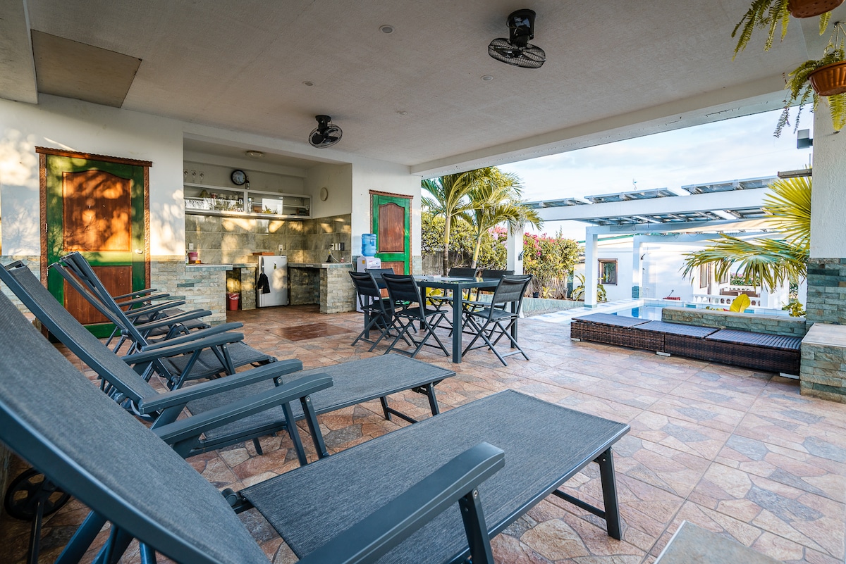 A shaded outdoor area is presented, featuring several lounge chairs arranged for relaxation. A dining table with chairs is visible nearby, alongside an outdoor kitchen setup. Lush greenery and a pool can be seen in the background, contributing to a serene atmosphere.