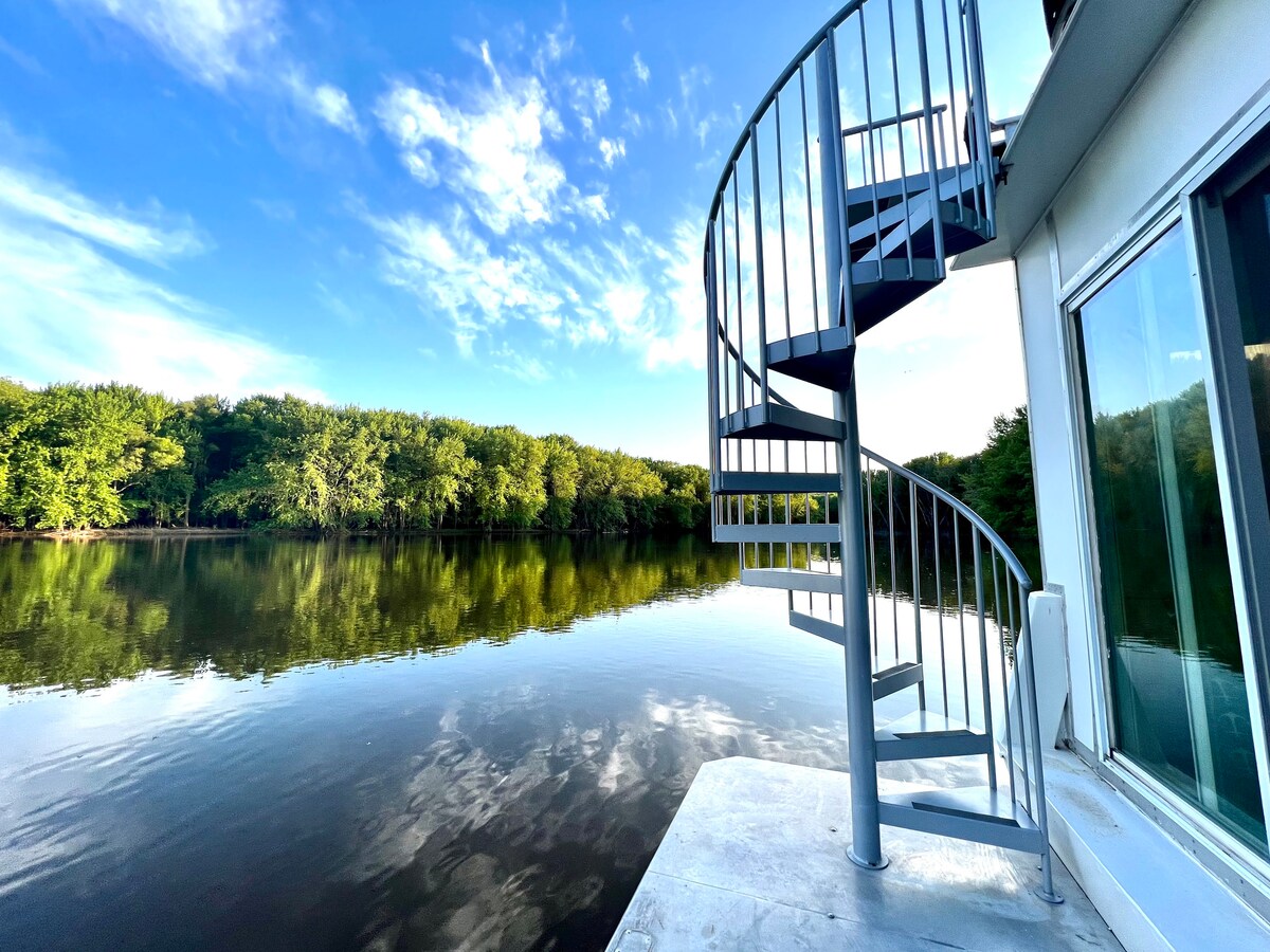 A spiral staircase leads from the houseboat to the roof, surrounded by tranquil waters reflecting a blue sky and lush green trees. The serene landscape evokes a sense of nature's beauty, offering a glimpse of the peaceful environment.