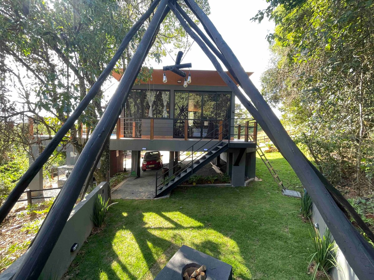 A contemporary chalet is framed by a triangular roof structure, surrounded by lush greenery. The outdoor area features a fire pit on a grassy lawn. A staircase leads to a covered balcony with large glass windows, providing a view of the outdoors.