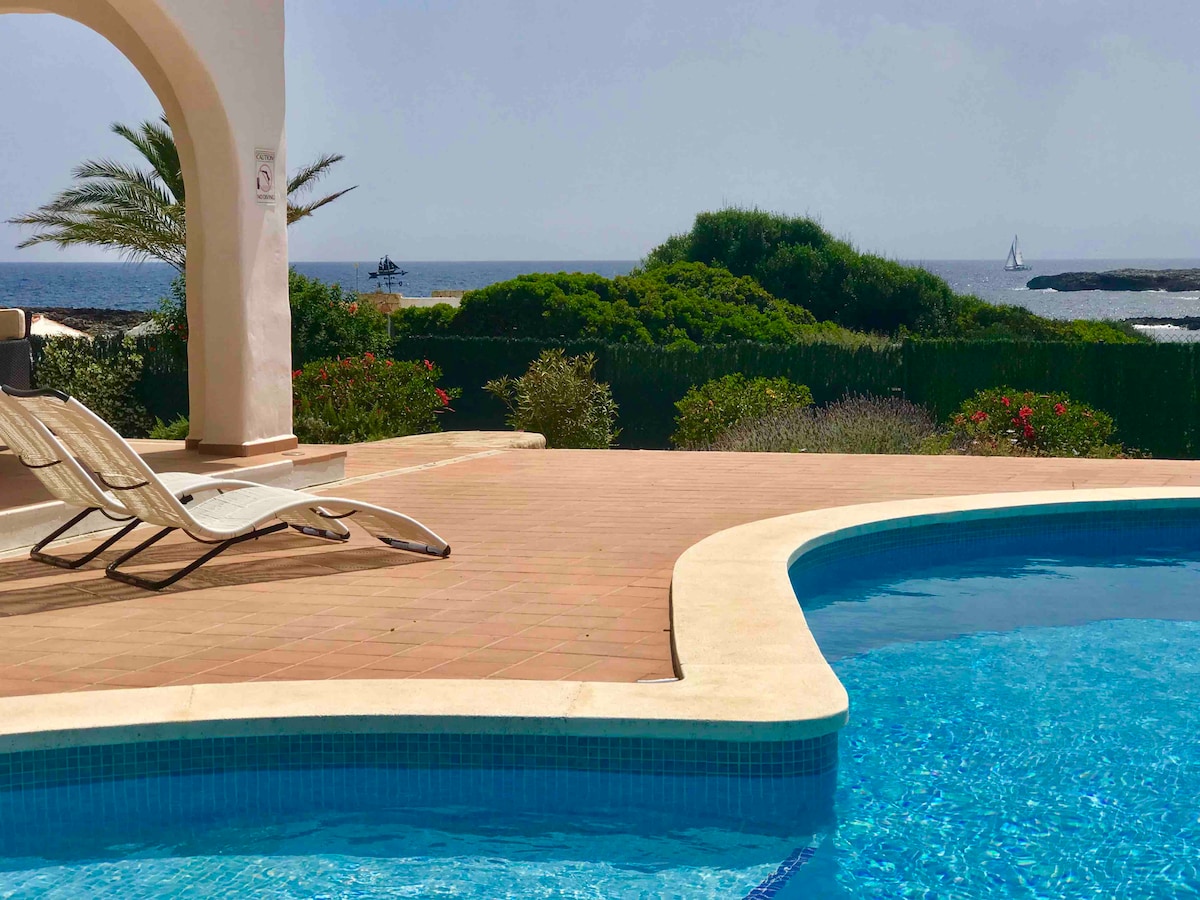 A spacious terrace features a clear salt-water pool reflecting sunlight. Comfortable lounge chairs are positioned nearby, while the shoreline and distant sailboats are visible in the background. Lush greenery frames the scene, enhancing the serene coastal atmosphere.