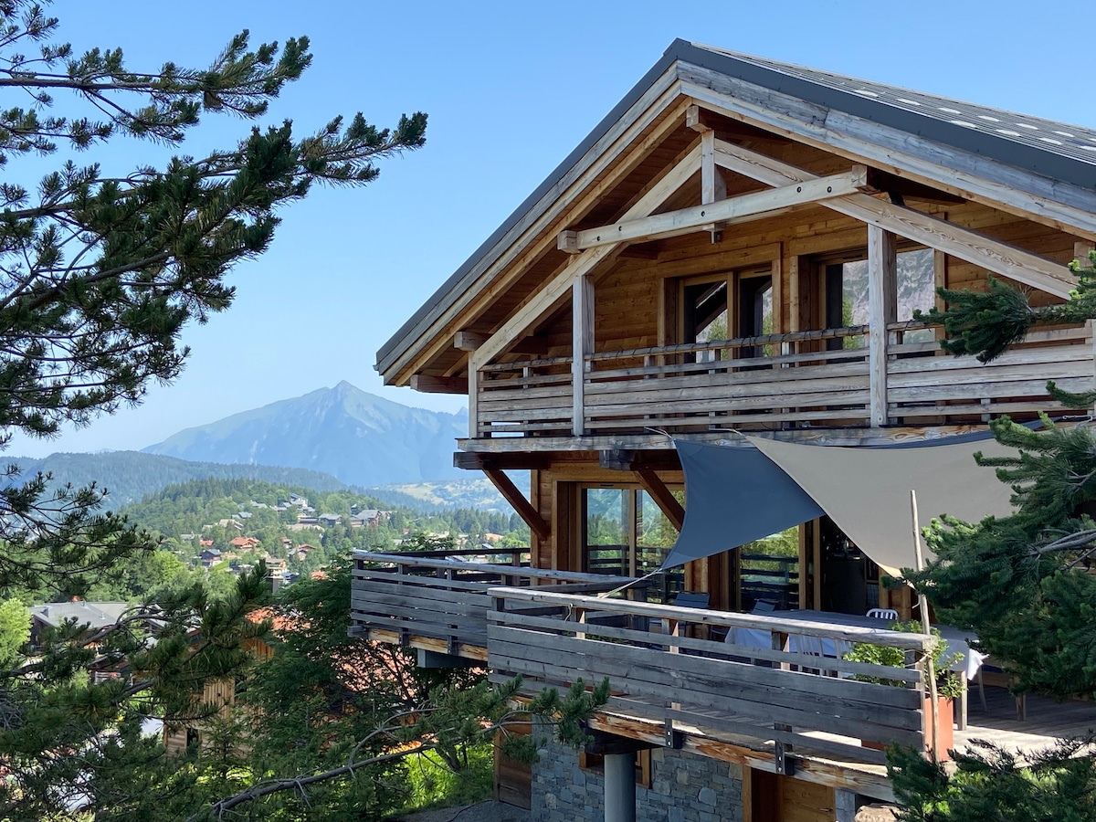 A modern chalet with wooden beams and large balconies is framed by lush greenery. The surrounding mountains are visible in the background, providing a serene setting. Sunlight reflects off the building, highlighting its natural materials and design.