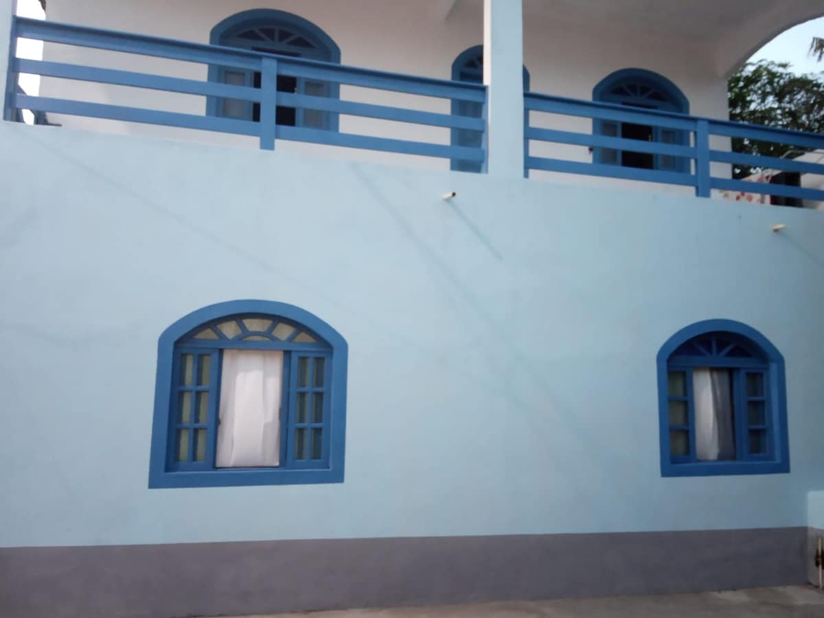 A light blue exterior wall features two arched windows with blue frames, each showcasing sheer white curtains. A balcony with blue railings is visible on the upper level, enhancing the building's charming design.