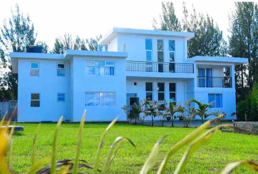A modern two-story house features a sleek white exterior and large windows. A balcony stretches across the second floor, overlooking a well-maintained green lawn dotted with shrubs. Tall trees frame the property, creating a sense of privacy and tranquility.