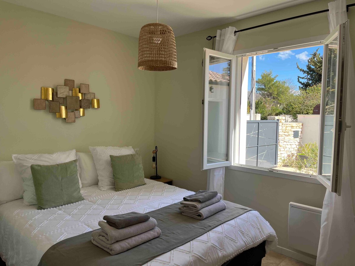 A cozy bedroom is presented with a spacious bed adorned with green accent pillows and a textured blanket. Natural light enters through an open window, illuminating the light-colored walls and a decorative wall piece. A nightstand is positioned beside the bed.