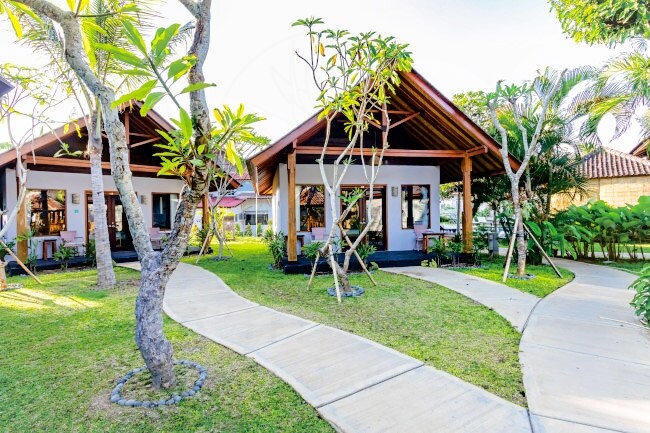 A scenic view of charming bungalow-style rooms set amidst lush greenery. A winding pathway made of light-colored concrete connects the bungalows, which feature sloping roofs and patio seating areas. Tropical trees and plants surround the space, creating a serene atmosphere.