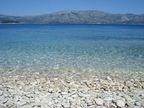 A clear blue sea meets a pebbled shoreline beneath a clear sky. The water transitions from deep blue to turquoise near the shore, revealing smooth, rounded stones of various sizes and colors. In the background, a mountainous landscape creates a serene backdrop.