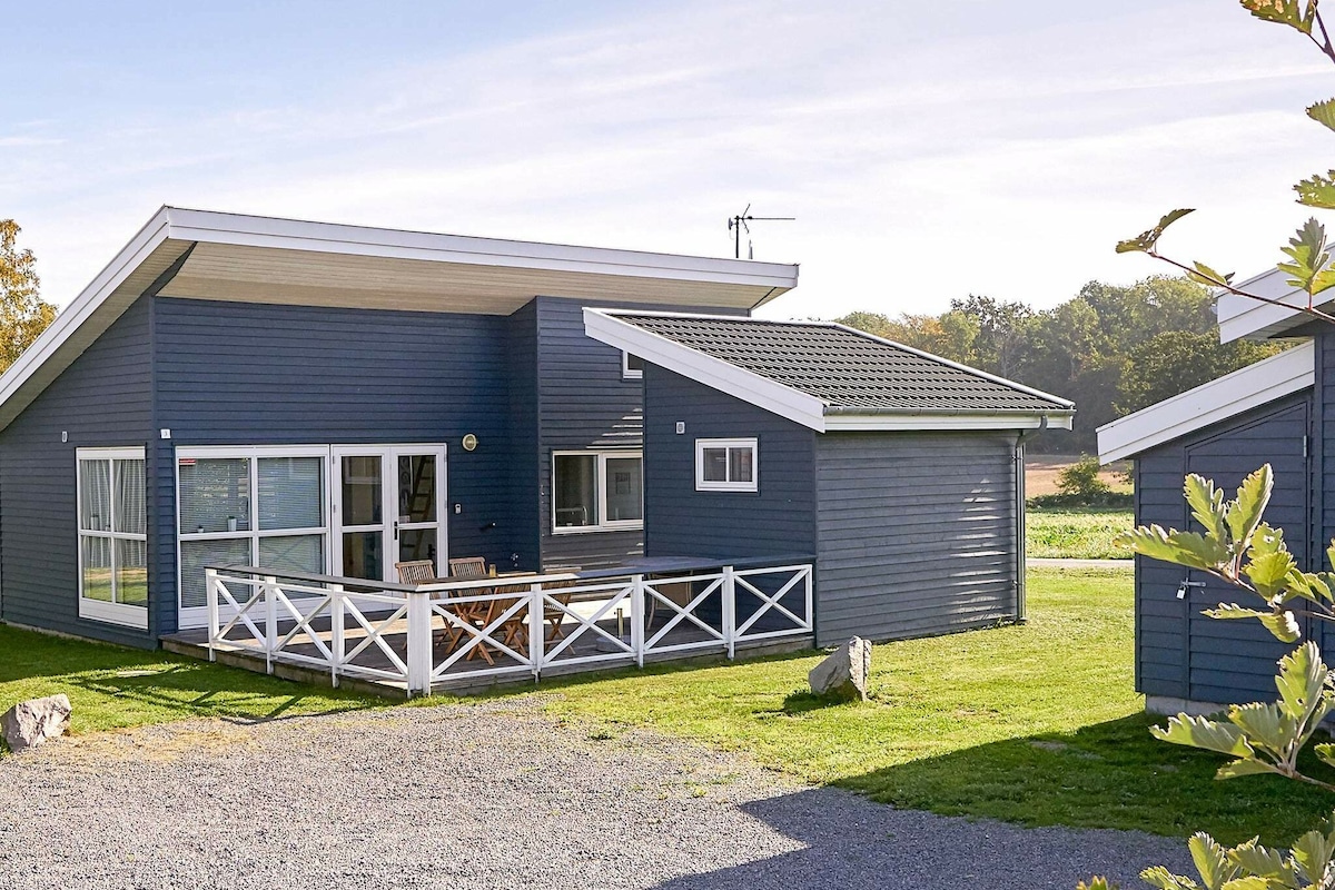 A modern holiday home is presented in a Scandinavian style, featuring large windows that invite natural light. A spacious terrace with wooden furniture extends from the house, surrounded by well-maintained green grass. The property includes an additional annex visible to the side.