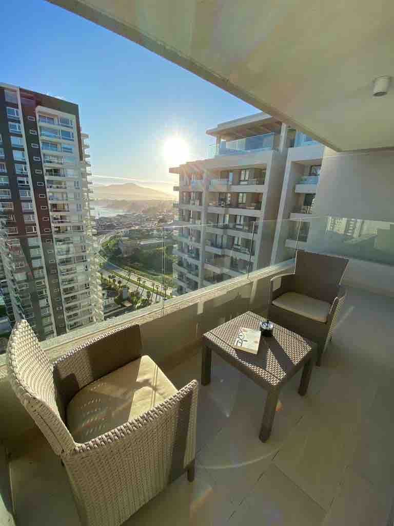 A private balcony is presented with two cushioned chairs and a small table, overlooking the stunning landscape. The sun rises in the background, illuminating the buildings and serene surroundings, providing a peaceful setting for enjoying mornings outdoors.