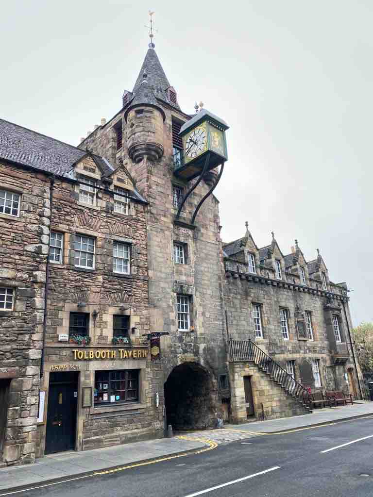 The historic Tolbooth Tavern is featured, showcasing its iconic architecture with a tall stone tower and detailed clock. The building's façade exhibits weathered stone and ornate features, while the surrounding street provides a glimpse of the bustling atmosphere in Old Town Edinburgh.