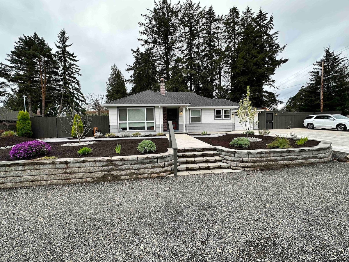 The exterior of the modern home is visible, featuring a manicured front yard with various plants and flowers. A stone pathway leads to the entrance, framed by low stone walls. Tall evergreen trees provide a natural backdrop, and ample parking is available in the gravel driveway.