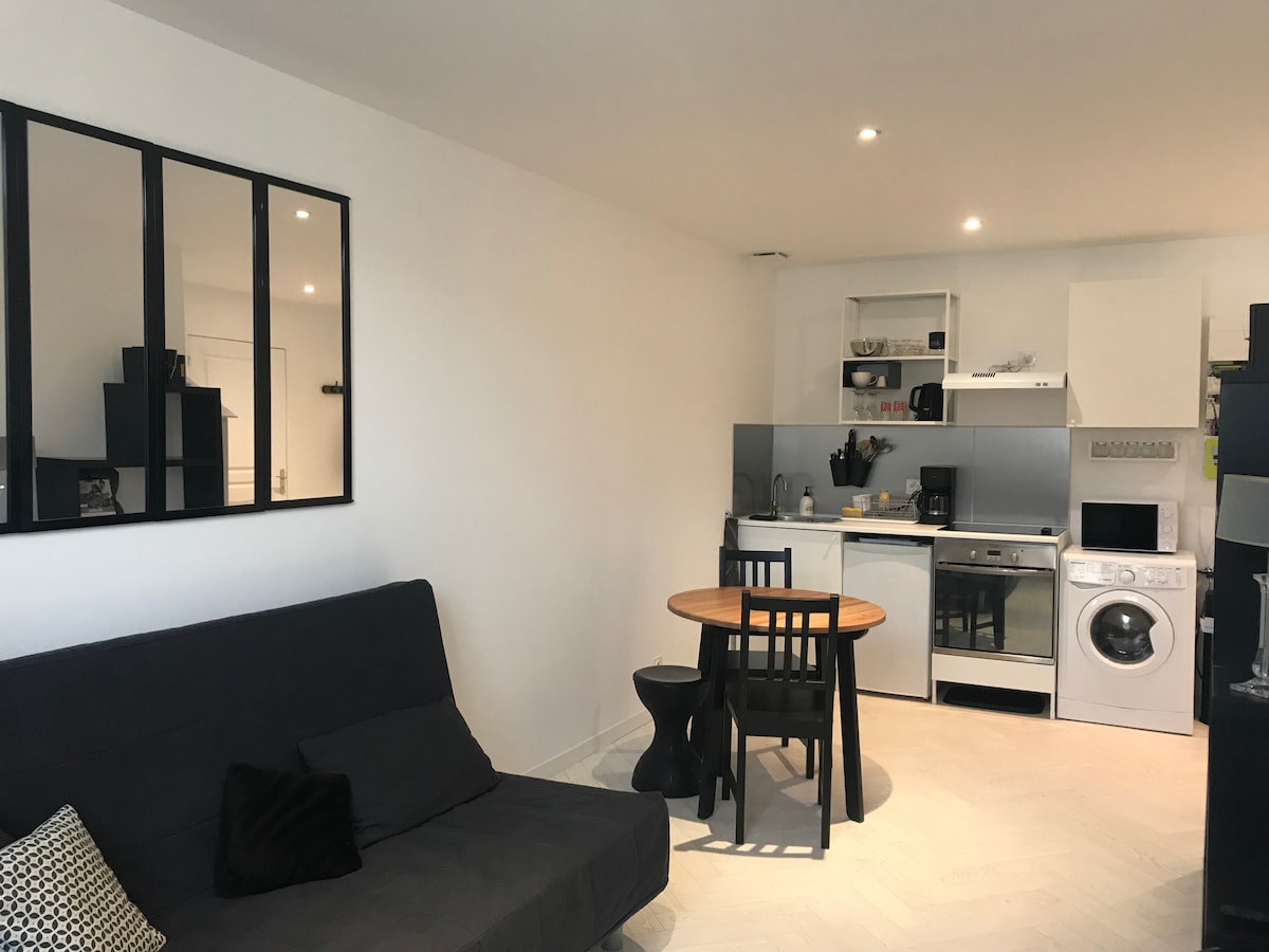 A modern living space is shown, featuring a comfortable sofa, a round dining table with two chairs, and a well-equipped kitchen area. Kitchen appliances are neatly arranged, and a washing machine is positioned alongside. Natural light brings a bright ambiance to the room.