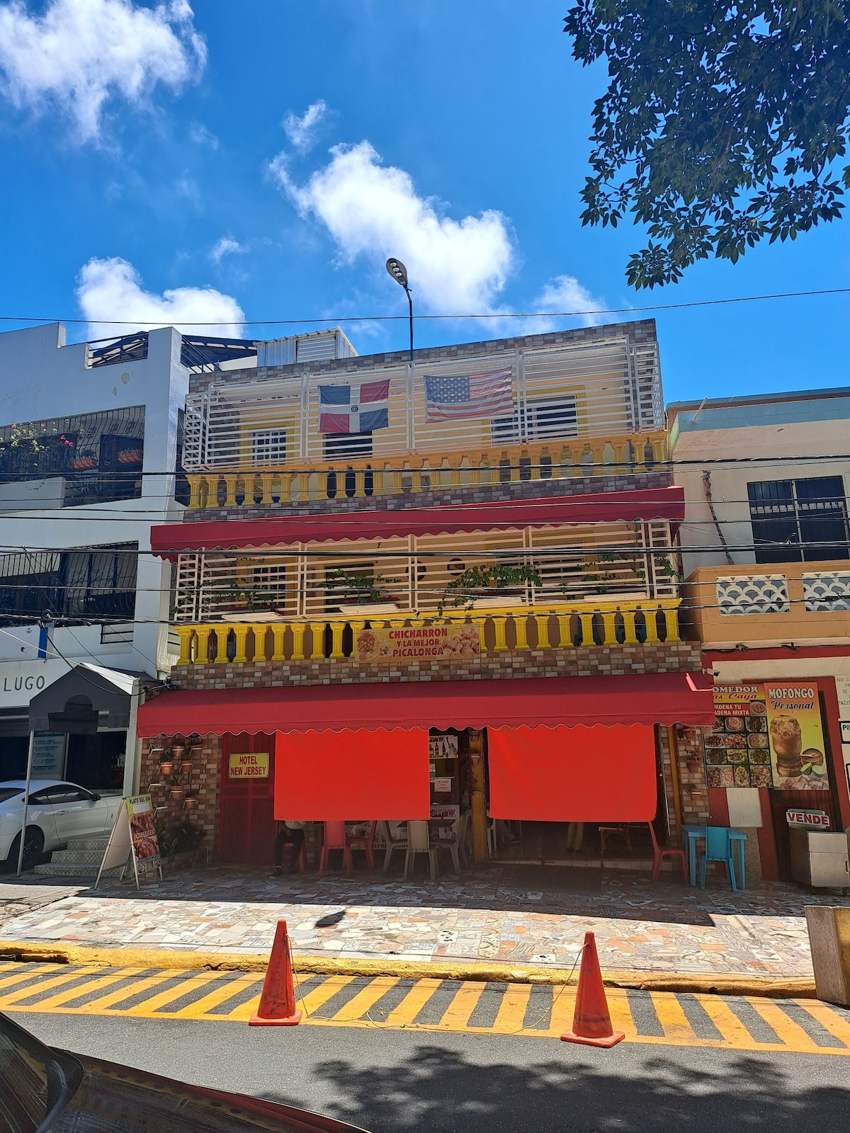 A vibrant multi-story building stands under a clear blue sky, adorned with colorful decorations and flags. A covered outdoor seating area is visible at the ground level, with several signs indicating nearby amenities. A yellow and black striped crosswalk runs in front of the property.