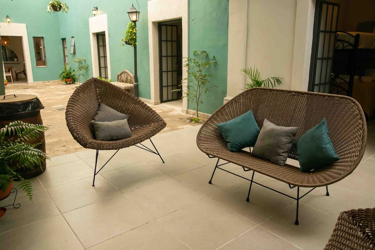 Two stylish wicker chairs are positioned around a central courtyard area, featuring soft cushions in various shades of teal. Light-colored tiles extend across the floor, while green plants add a touch of nature to the inviting space.