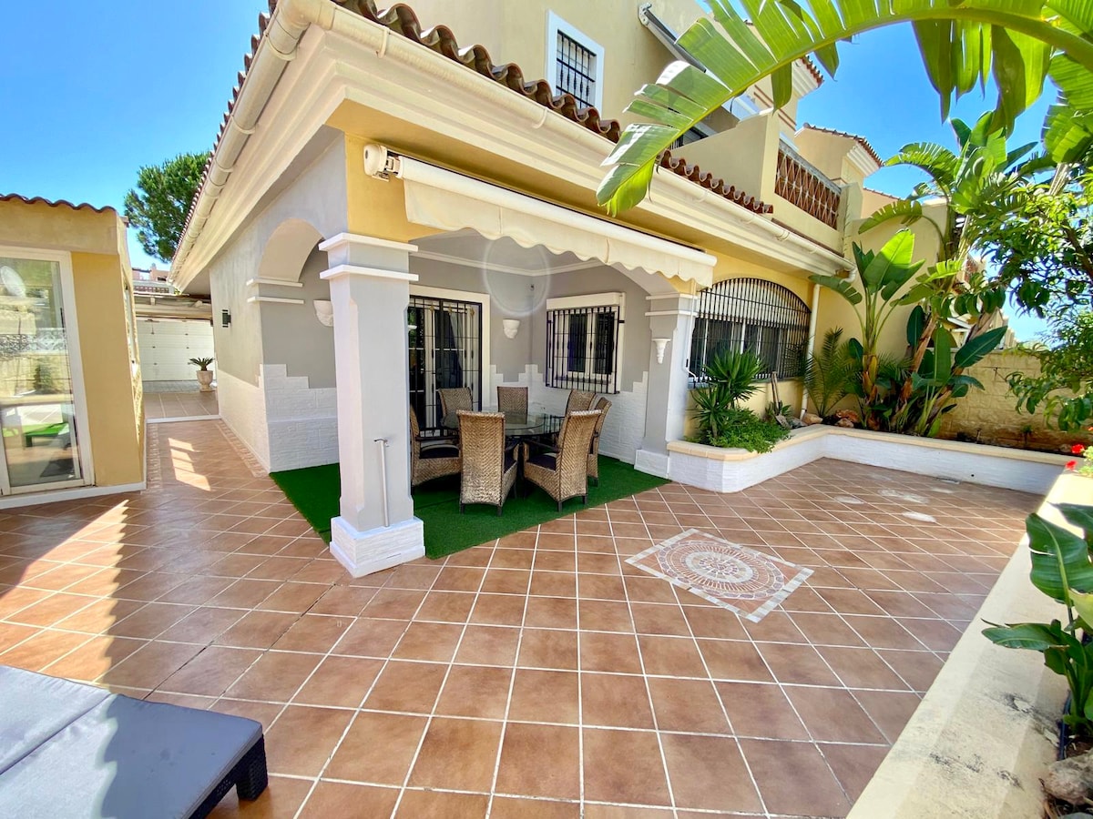 An outdoor terrace is presented, featuring a tiled floor with a decorative design at its center. Comfortable seating arrangements made from natural materials are surrounded by lush plants, creating a cozy space for relaxation. Sunlight illuminates the area, enhancing its inviting atmosphere.
