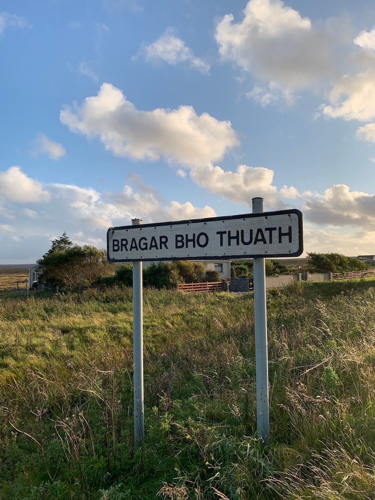 A roadside sign indicating 'Bragar Bho Thuath' is observed, framed by lush green grass and a clear sky with scattered clouds. The sign stands prominently, pointing toward the village, with hints of structures visible in the background.