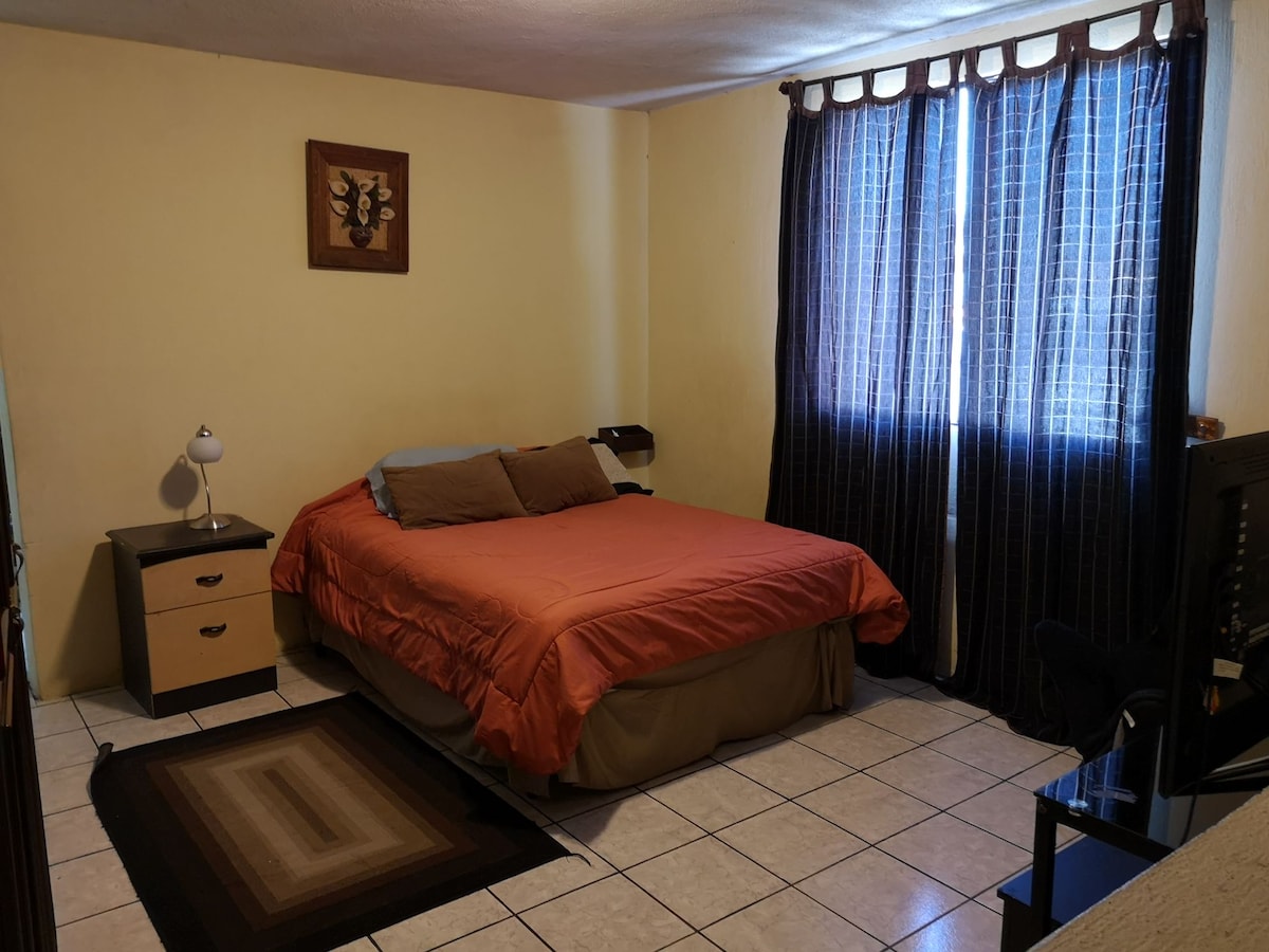 A private bedroom features a spacious bed with orange and brown bedding. A nightstand with a lamp sits beside the bed, and a dark curtain beautifully frames the window. A television and a small chair are visible across the room, enhancing the functional space.