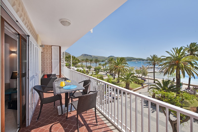 A spacious terrace is presented, featuring a table set for outdoor dining, accompanied by four wicker chairs. Lush palm trees and the beach can be seen below, with a view of the ocean and coastal landscape extending beyond. The sky is clear and sunny.