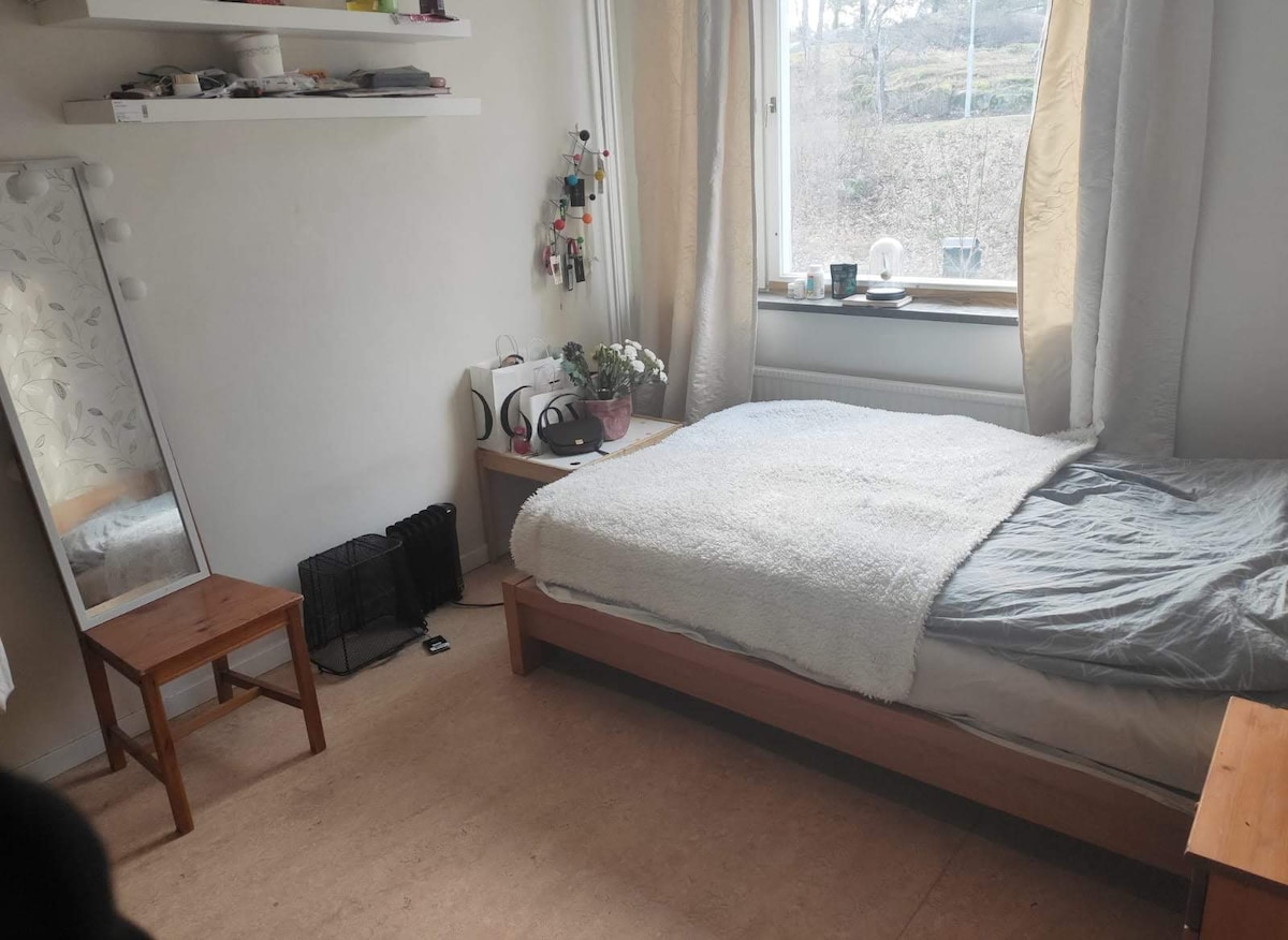A bright bedroom features a spacious bed with a light gray blanket, accompanied by a bedside table with a kettle and potted plants. A full-length mirror leans against the wall, reflecting sunlight from the window. Shelves above the bed display various items.
