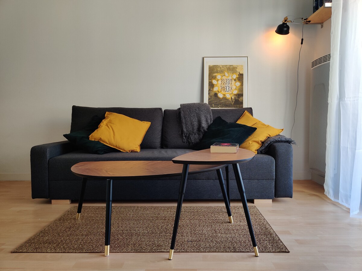 A modern living area features a dark grey sofa adorned with yellow and green cushions. A unique coffee table with a wood top and slender black legs sits on a textured rug. Natural light is invited through a nearby window, highlighting the warm tones of the space.