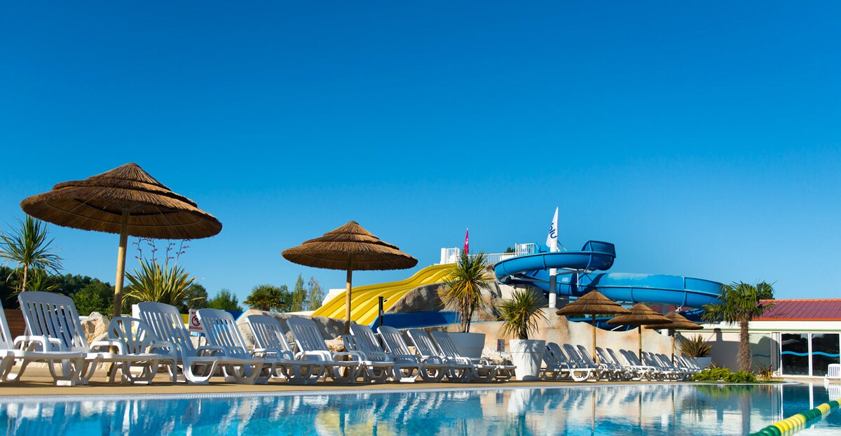 A sunny outdoor pool area is visible, featuring lounge chairs lined up beside the water. Thatched umbrellas provide shade. A water slide and colorful play structures are situated in the background, under a clear blue sky.