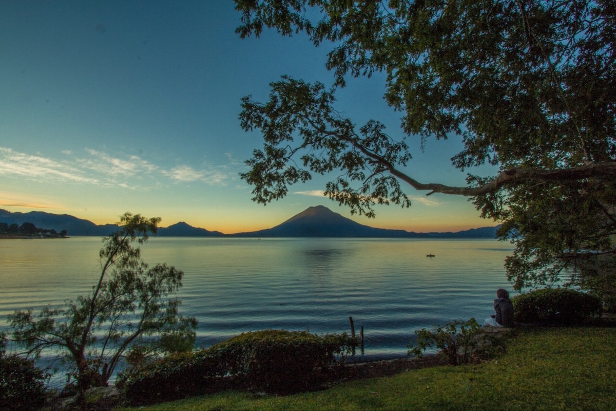 A serene view of the lake is captured at sunset, with gentle waves reflecting the colors of the sky. The surrounding greenery frames the scene, while distant mountains rise majestically in the background, enhancing the tranquility of the environment.
