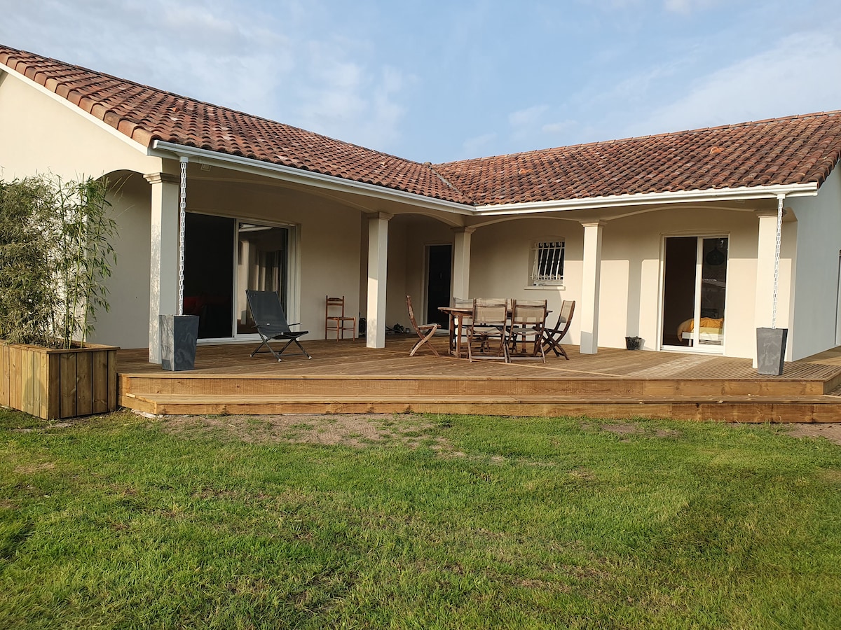 A single-story house is shown, featuring a spacious wooden deck surrounded by a grassy area. Outdoor seating options are visible, including a dining table and lounge chair. Large glass doors provide access to the interior, enhancing the connection to the outdoor space.