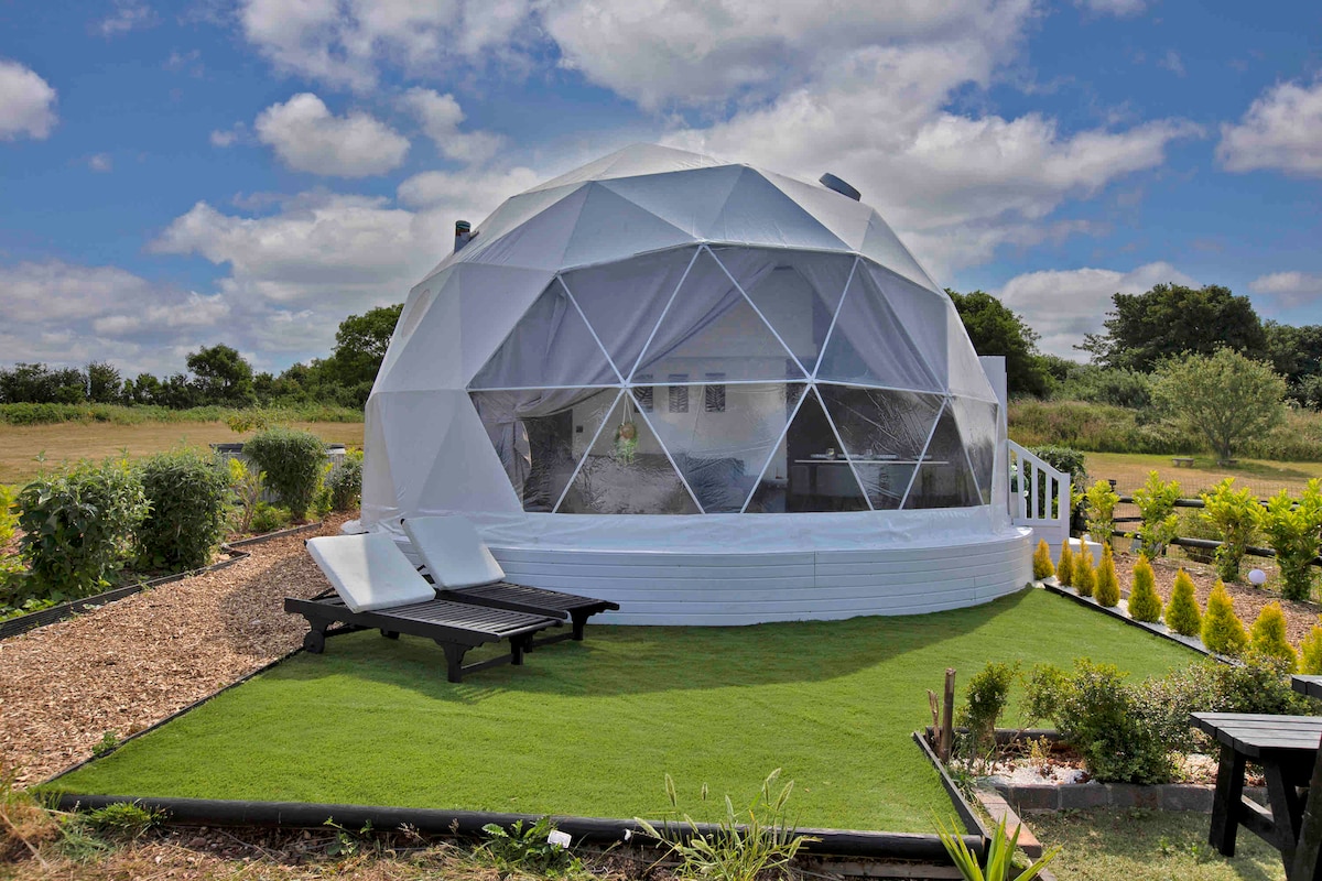 An eco-friendly dome features a geodesic design, surrounded by a verdant garden. The exterior is composed of white panels, with large transparent windows allowing natural light to flood the interior. Sun loungers are positioned on groomed grass, providing a tranquil outdoor relaxation area.
